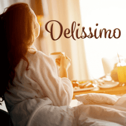 Darkhaired woman in a bathrobe holds a cup and looks at the window, on the front of the photo is the logotype: Delissimo